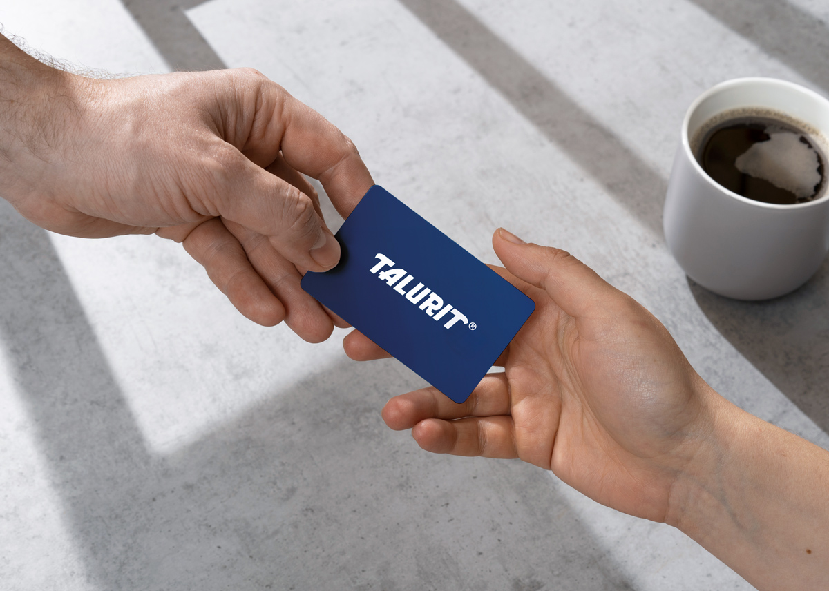 One hand is passing a blue card with the white TALURIT logo to another hand. A white coffee cup sits on a gray surface in the background. © Copyright Talurit AB. All rights reserved.