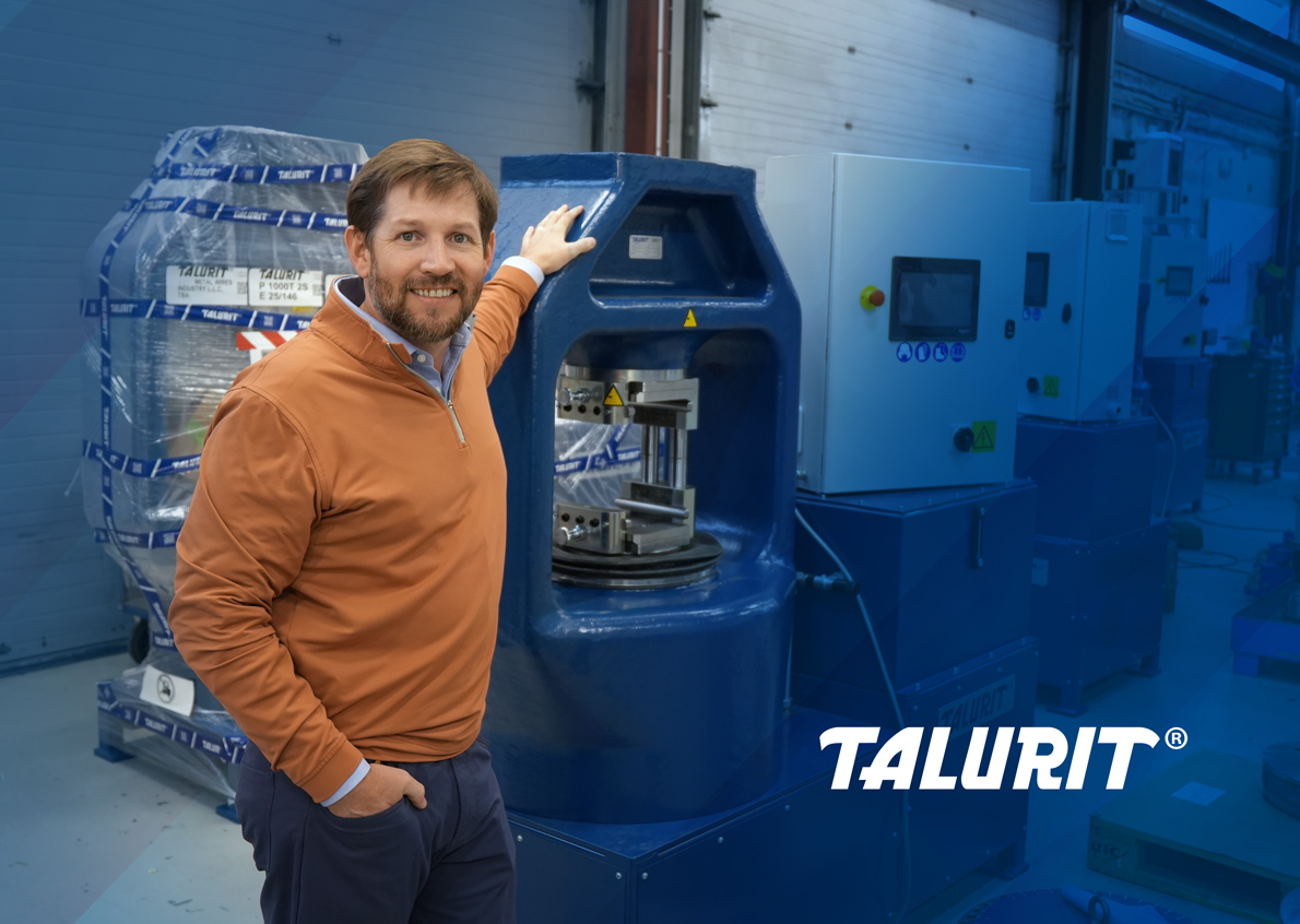 Luke-Staten-Vice-President-at-Talurit-Inc_ED_1190px Talurit® Luke Staten, wearing an orange sweater and blue pants, stands beside a large blue hydraulic swager in a factory setting. The word "TALURIT" is displayed in the lower right corner. © Copyright Talurit AB. All rights reserved.