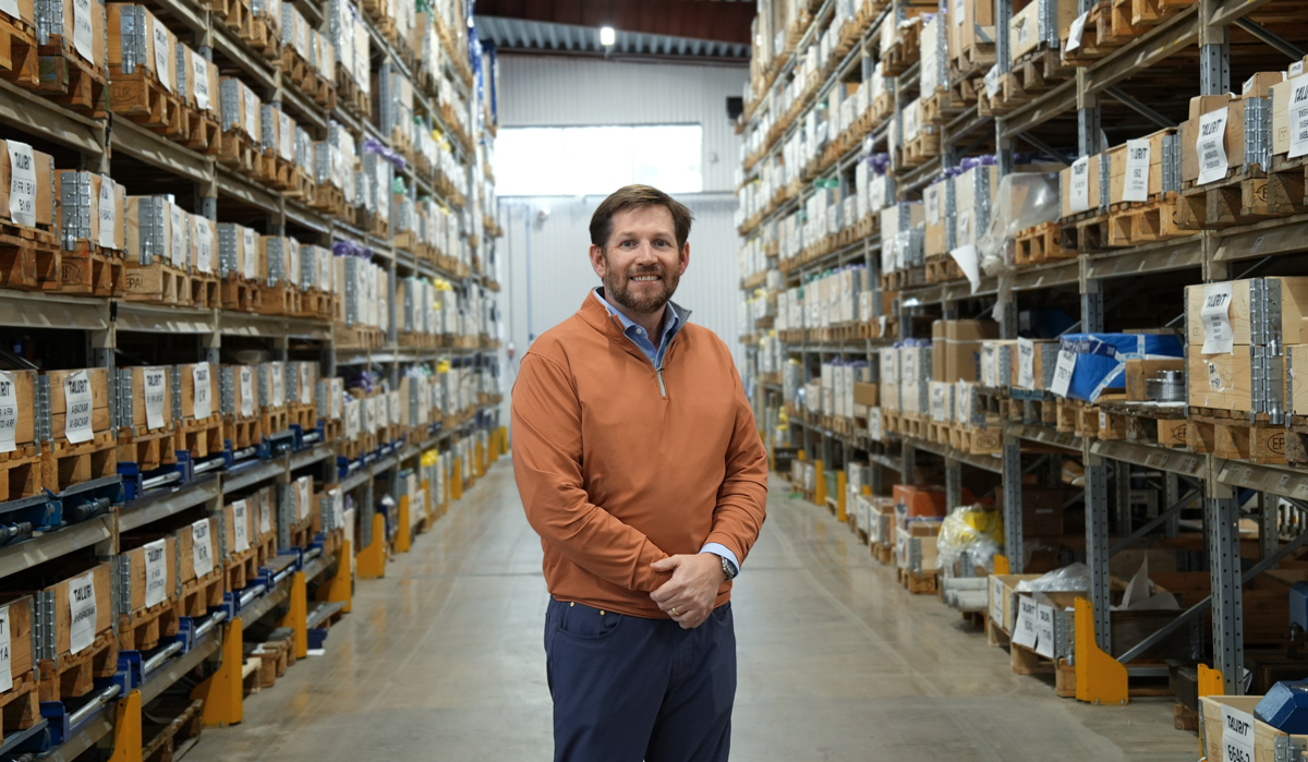 Talurit® Luke Staten stands in the aisle of a warehouse lined with shelves full of organized boxes and packages. © Copyright Talurit AB. All rights reserved.